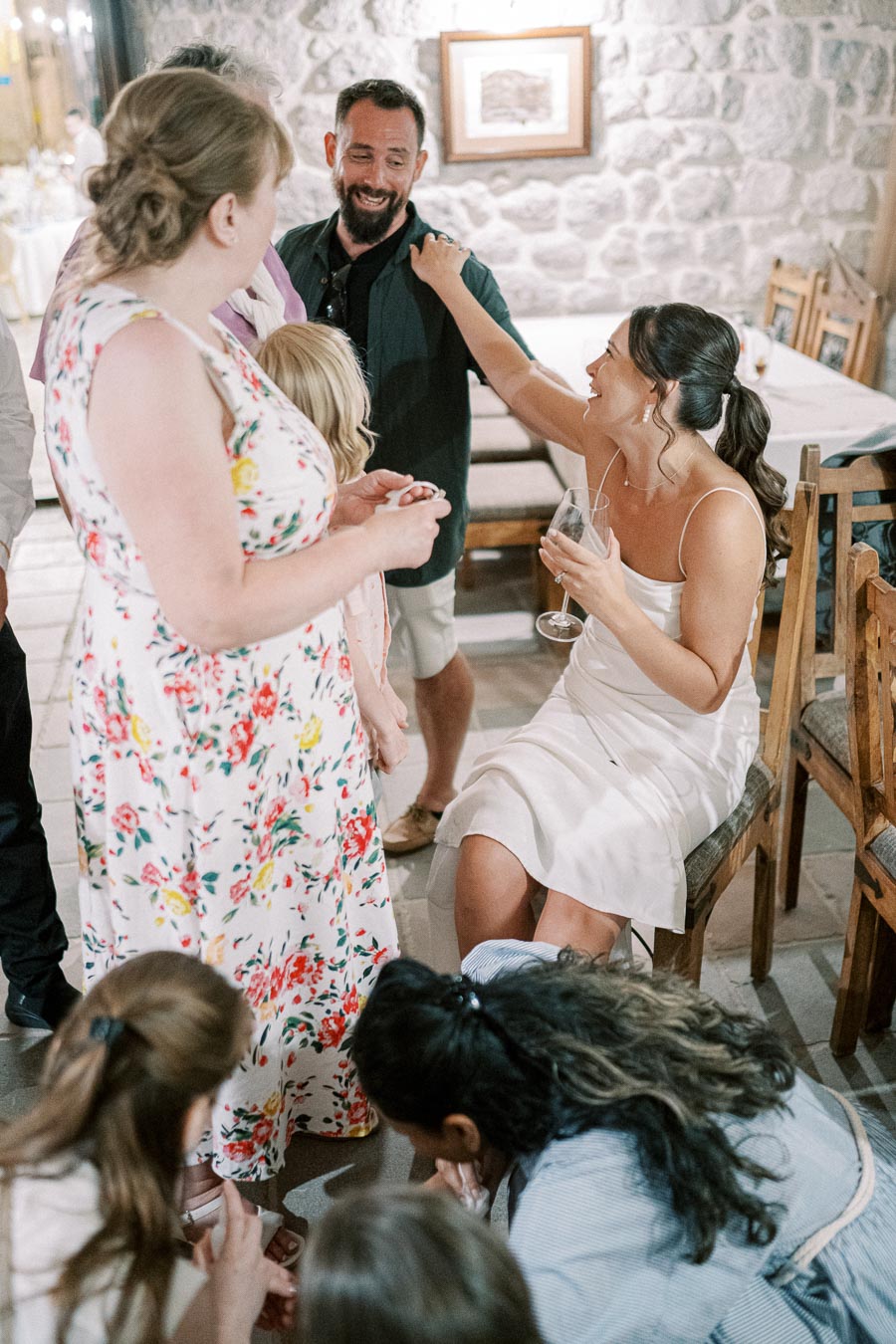 Group of people enjoying a lively gathering, with a woman in a white dress smiling and holding a wine glass, surrounded by friends in casual attire, set in a rustic stone room.