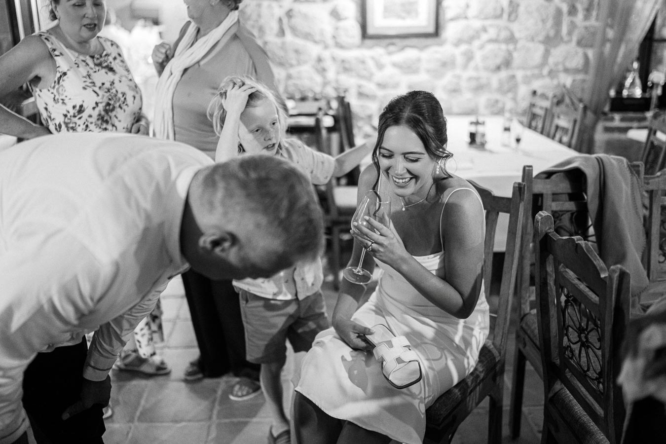 A joyful group of people gathered in a cozy indoor setting, with a woman in a white dress laughing and holding a wine glass, surrounded by smiling individuals engaged in lively conversation.