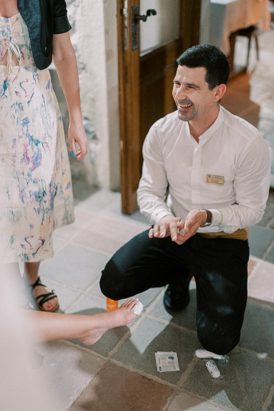 A man in a white shirt kneels on a stone floor, smiling as he bandages a woman's foot. Another woman in a floral dress stands nearby, offering assistance. The scene suggests a first aid moment, possibly at an event or gathering.