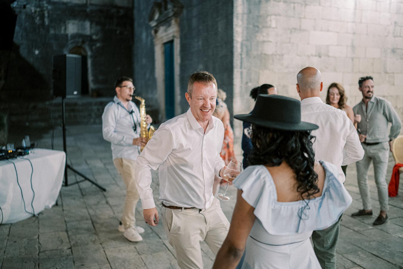 A lively group of people enjoying a night out, with a saxophonist playing music, and individuals dancing and socializing in an outdoor setting with stone walls.