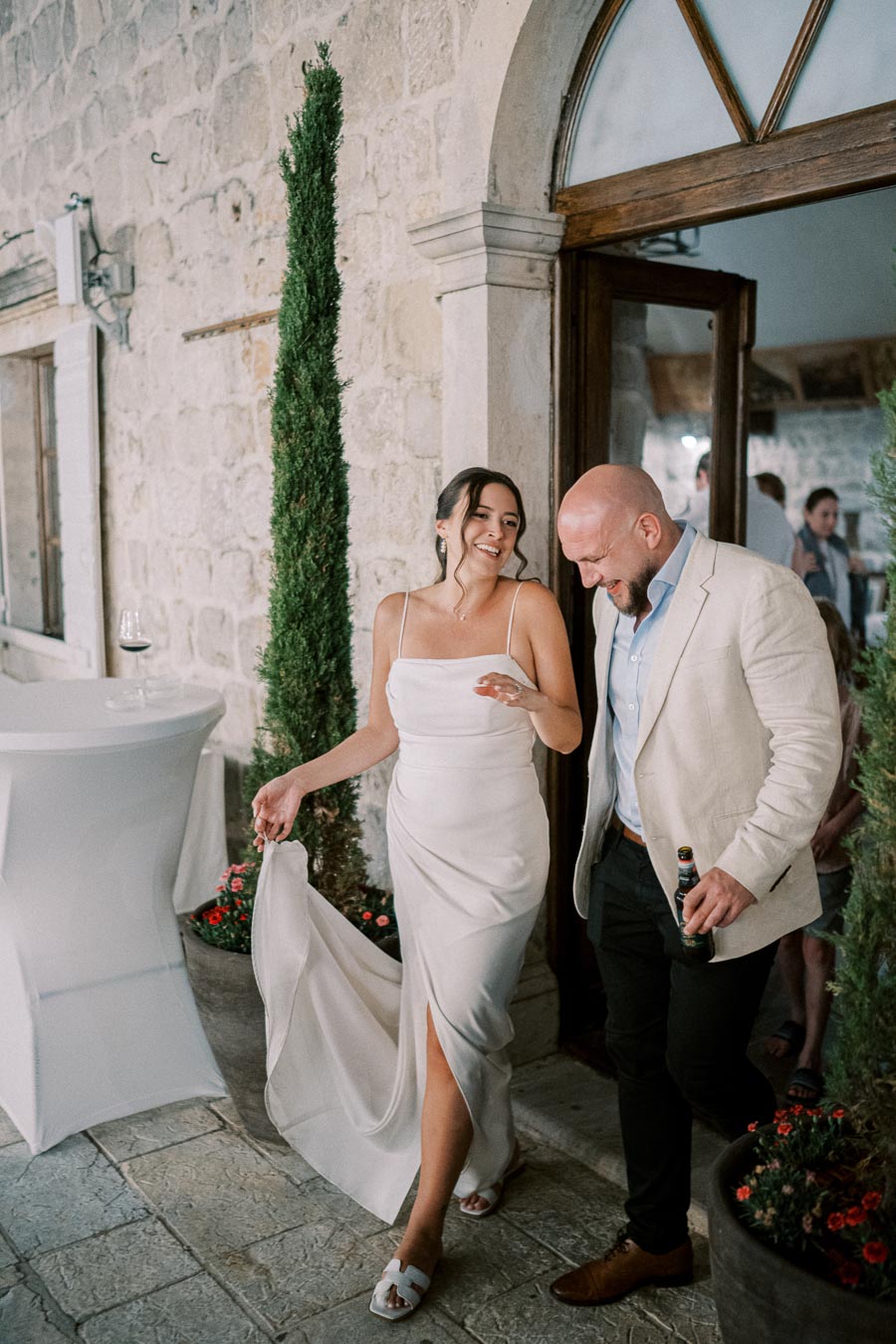 A smiling woman in a white dress and sandals walking with a man in a light blazer, enjoying an outdoor event by a stone building.