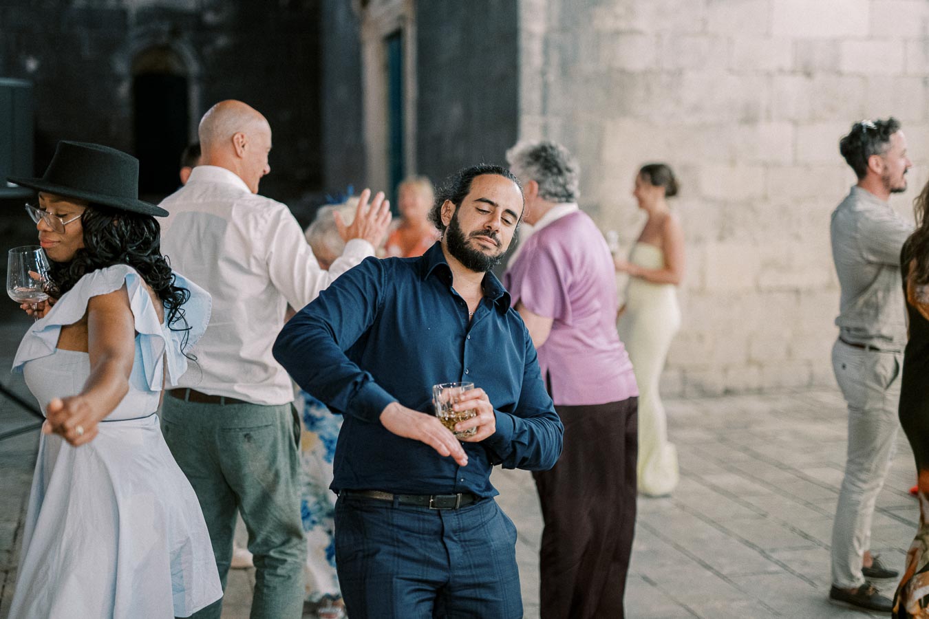 A group of people dancing at an indoor event, with one man in a blue shirt holding a drink and enjoying the music.