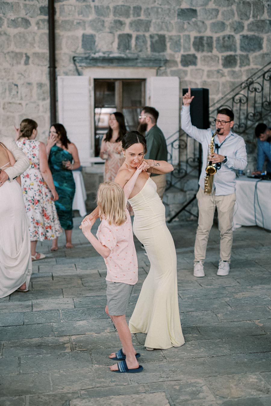 A woman in a yellow dress and a young boy dance together at an outdoor event, with a group of people socializing in the background. A musician plays a saxophone near a stone building with a window.