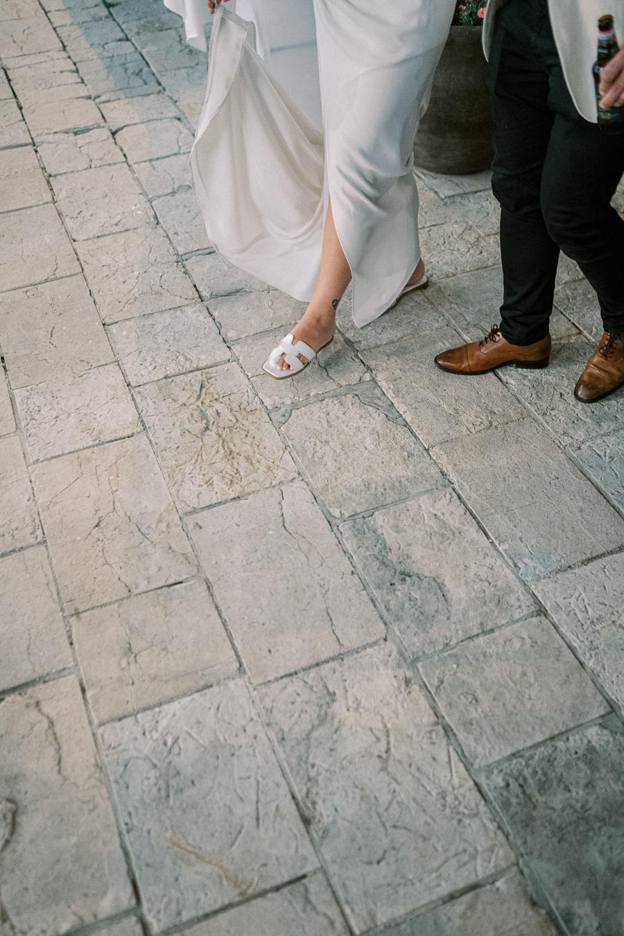 Close-up of a bride in a flowing white dress and sandals walking on a stone pavement alongside a man in formal shoes, capturing a candid wedding moment.