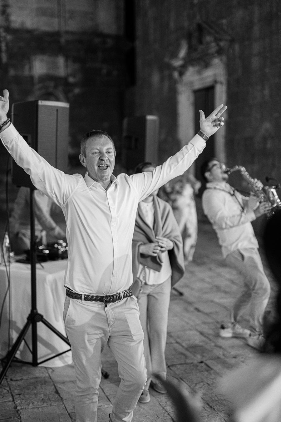 A black and white photo capturing a lively celebration, featuring a joyous man in a white shirt with arms raised, accompanied by a saxophonist performing energetically, with blurred guests in the background.