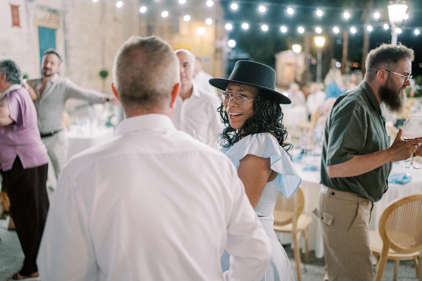 A joyful evening gathering with people socializing under string lights, featuring a woman in a stylish hat and glasses smiling and engaging in conversation.