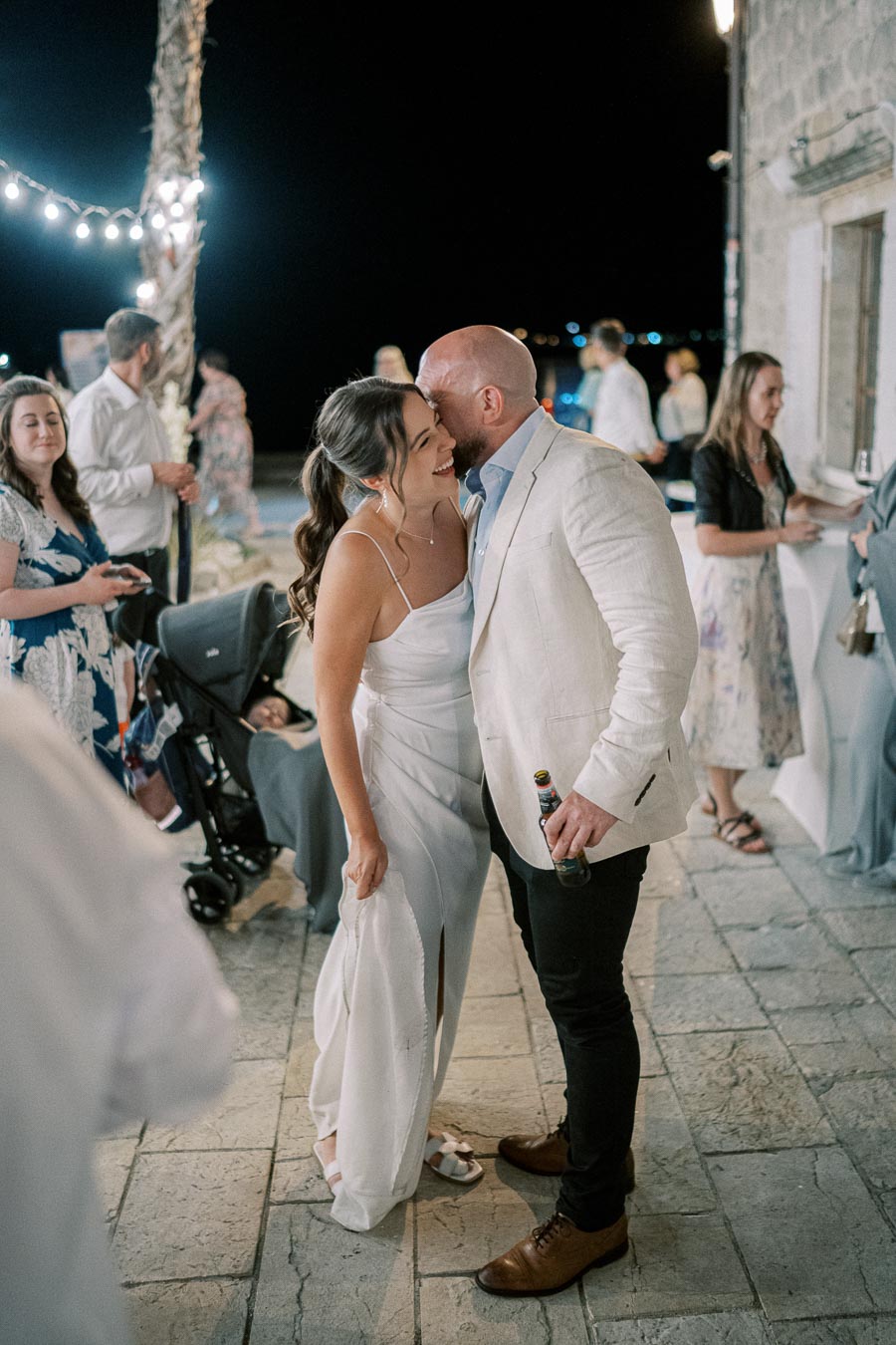 A couple sharing a joyful moment at a nighttime outdoor wedding reception, surrounded by warmly lit decor and elegantly dressed guests.