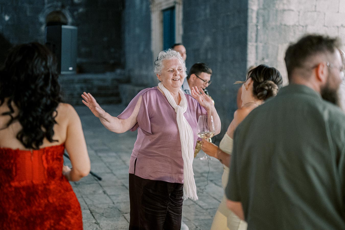 Elderly woman dancing joyfully at a party with other guests, wearing a purple blouse and white scarf, holding a glass of wine, while engaging in lively conversation and celebration