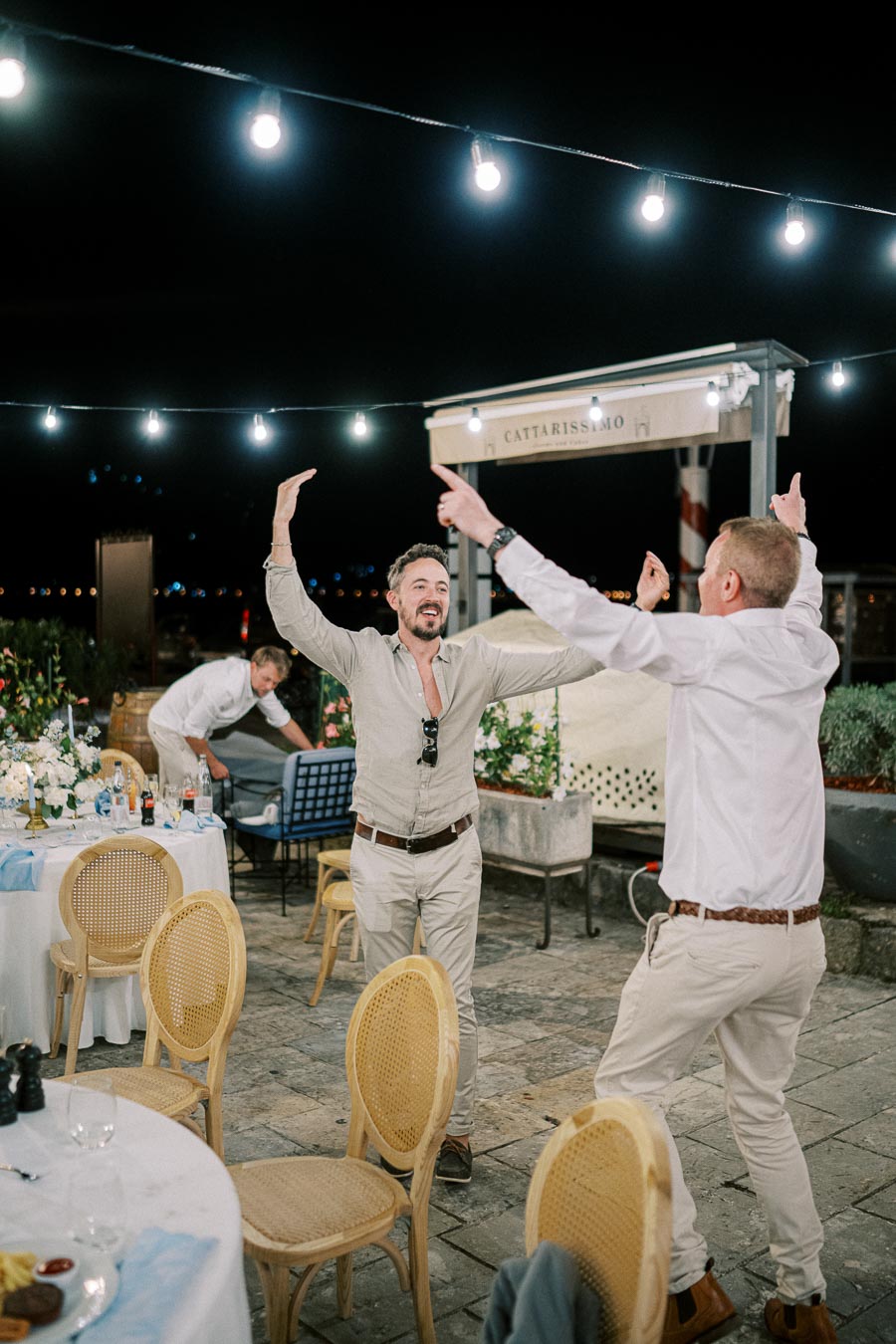 Men celebrating at an outdoor evening event, surrounded by decorated tables and string lights, enjoying a festive atmosphere.