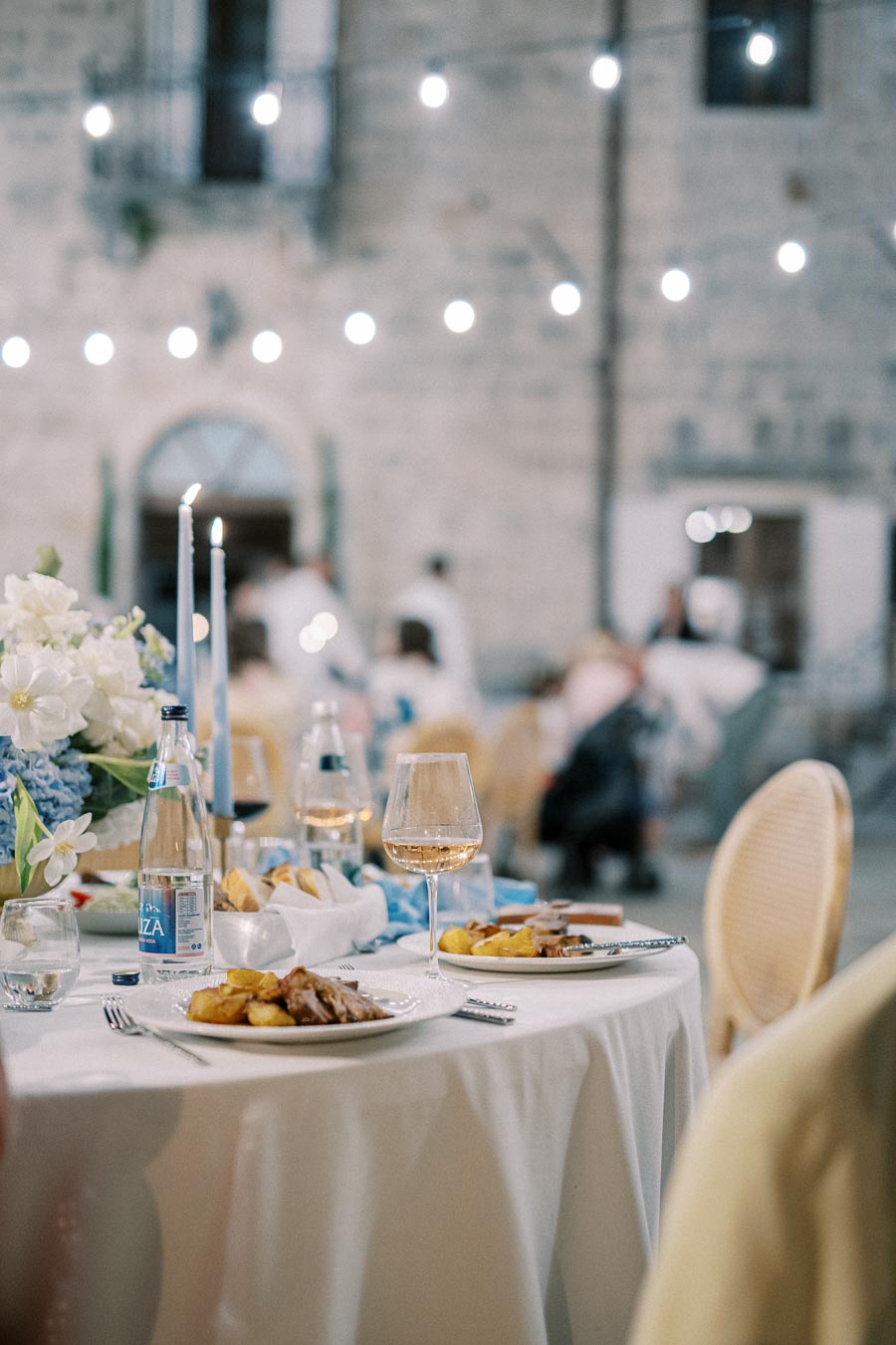 Elegant outdoor dining setup with a focus on a table featuring plates of food, glass of wine, bottled water, and floral centerpiece under string lights in a rustic courtyard setting.