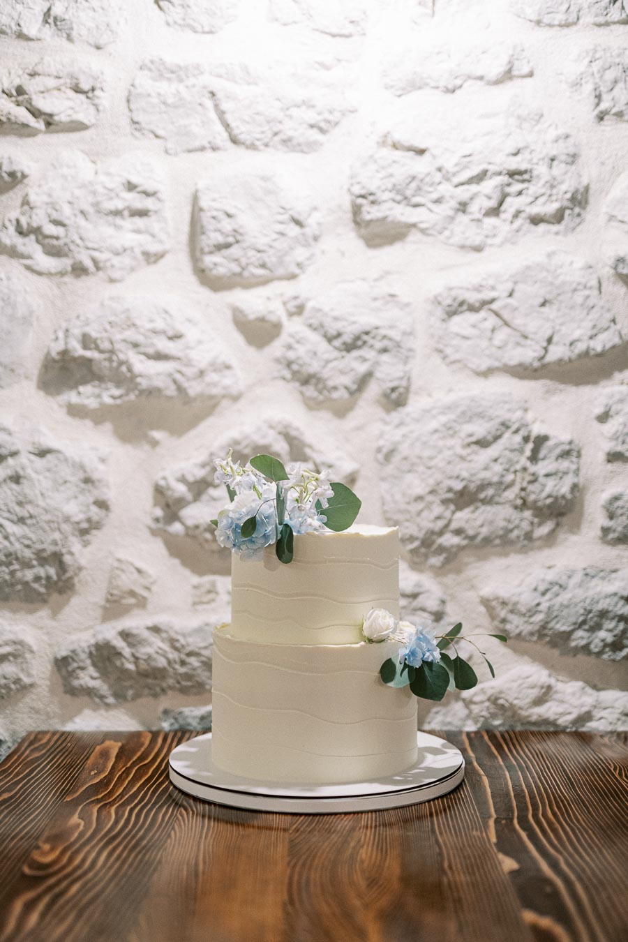 Two-tier white wedding cake with elegant floral decorations in blue and white, set against a rustic stone wall background.