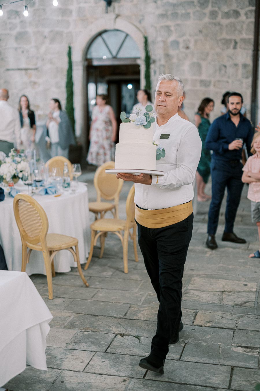 A waiter carrying a tiered wedding cake adorned with floral decorations at an elegant outdoor reception, with guests in the background and tables set with white tablecloths and blue accents.
