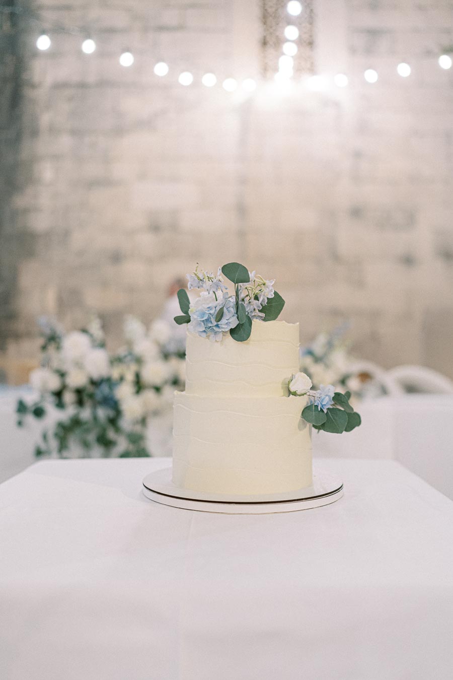 A two-tiered white wedding cake adorned with delicate blue and white flowers, set on a table in a softly lit rustic venue.
