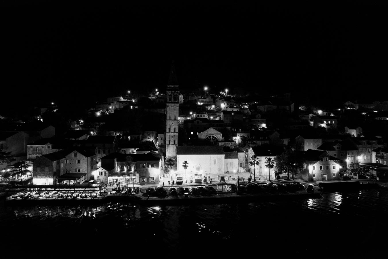 Black and white nighttime view of a coastal town with illuminated buildings, a prominent church bell tower, and reflections on the water.
