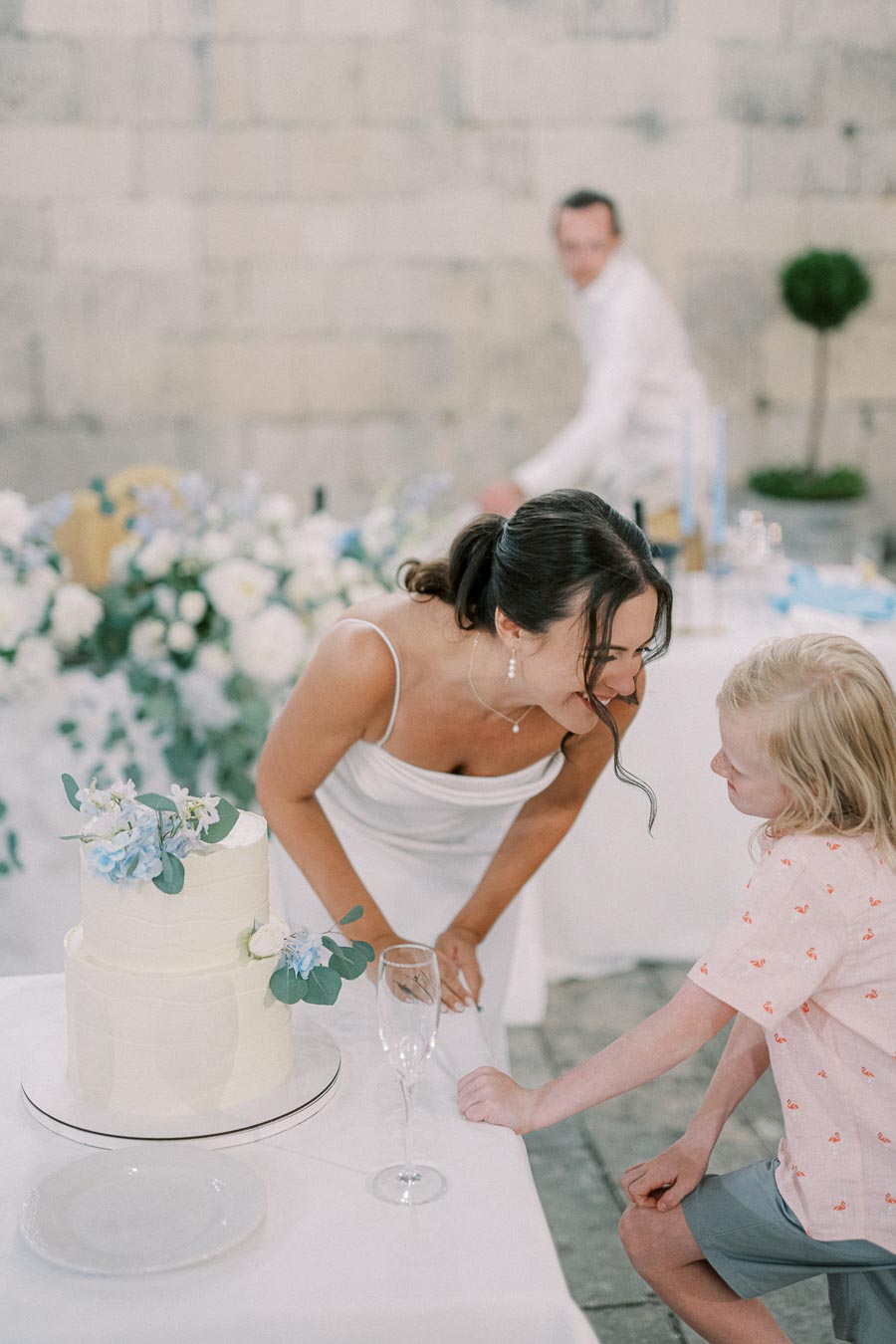 Woman in white dress smiling at child beside a decorated wedding cake, with elegant floral arrangements and champagne glass in elegant setting.
