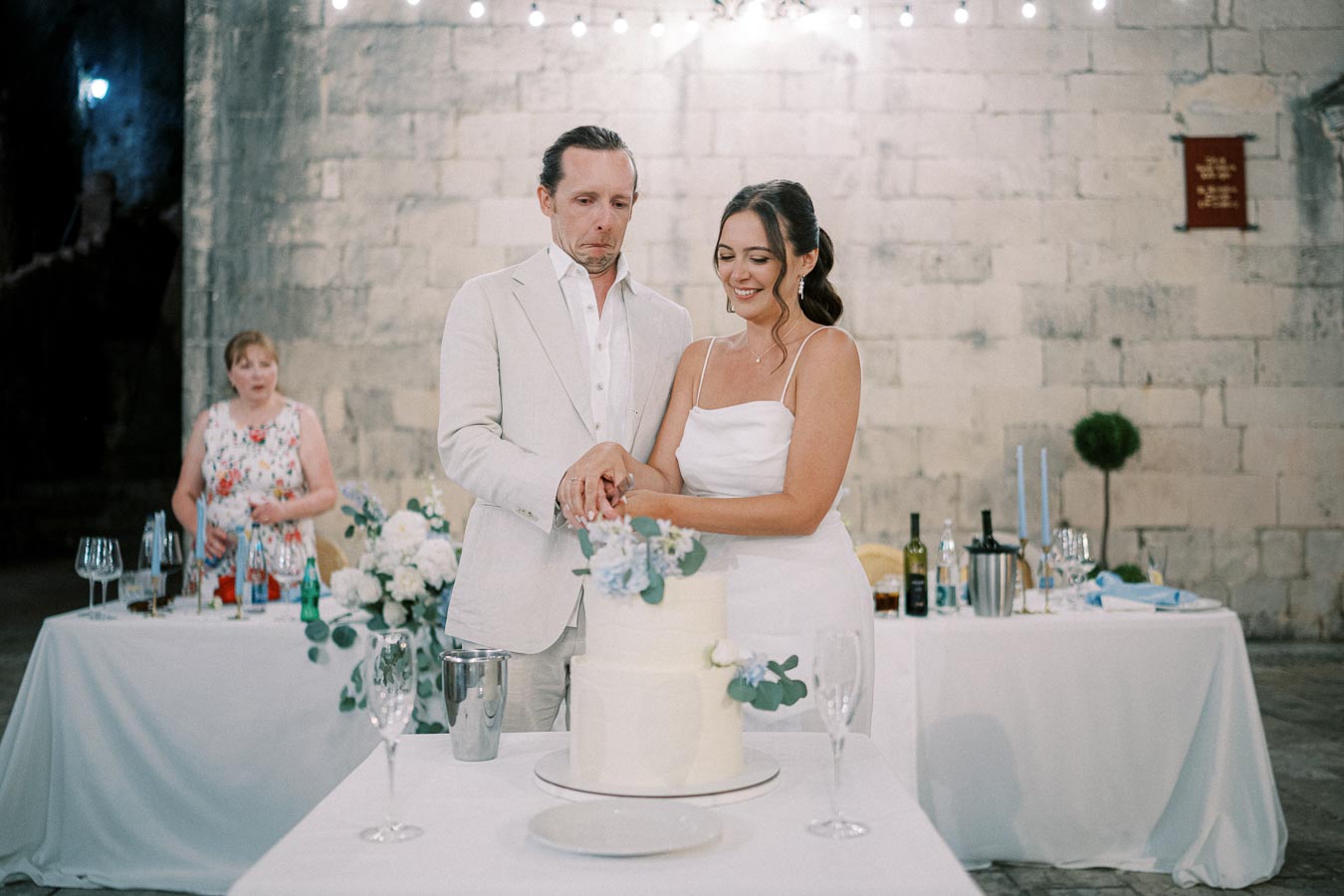 Elegant bride and groom cutting a white wedding cake adorned with blue flowers in a rustic, softly lit reception setting, surrounded by decorative tables and floral arrangements.