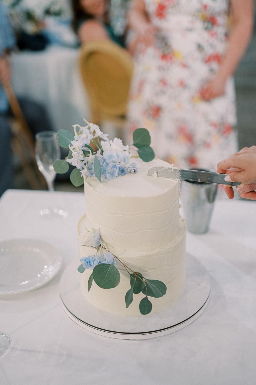 Two-tier white wedding cake adorned with blue and white flowers, being sliced at a wedding reception.