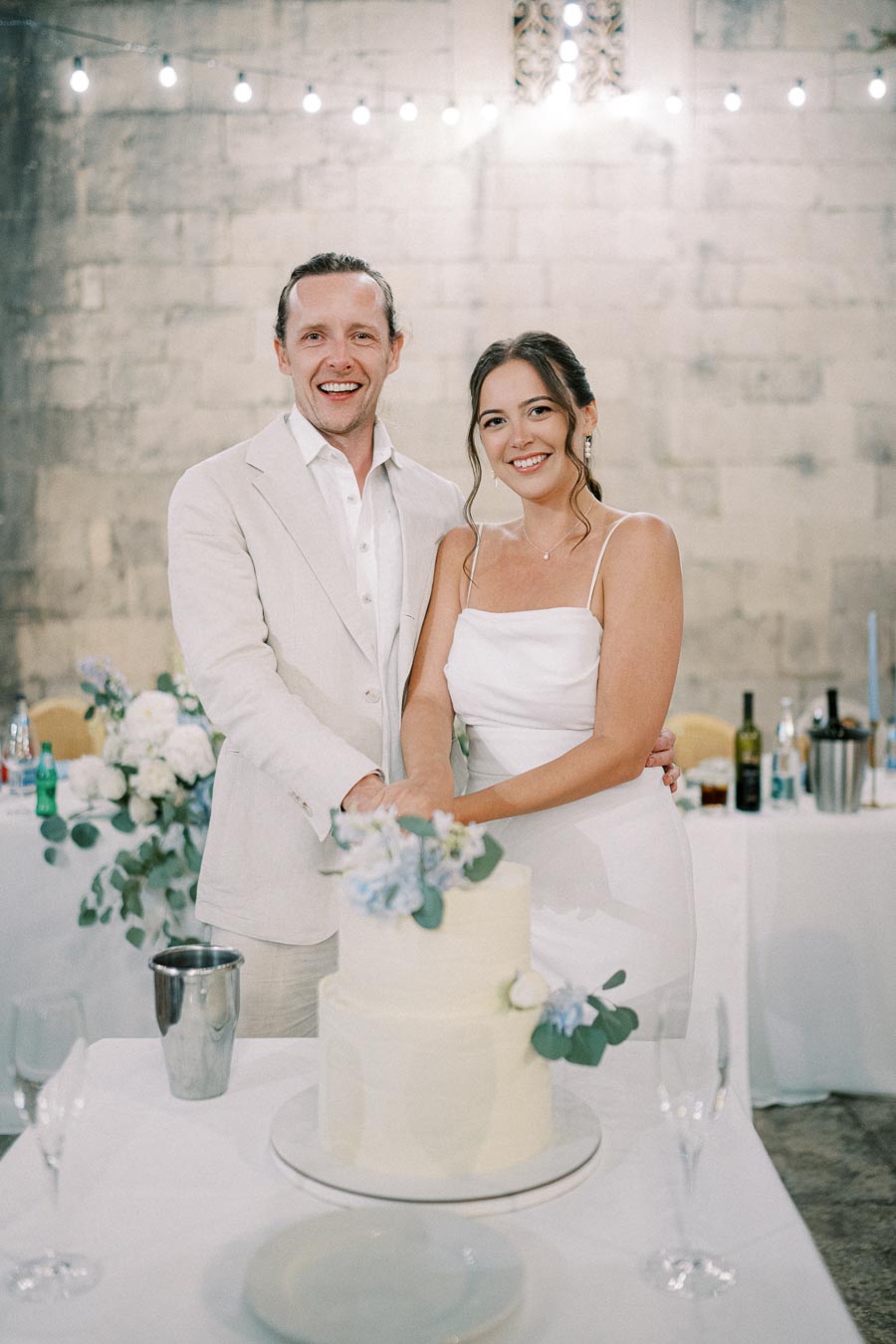 A happy couple in elegant attire joyfully cutting their wedding cake, topped with delicate floral decorations, set in a beautifully lit reception venue.
