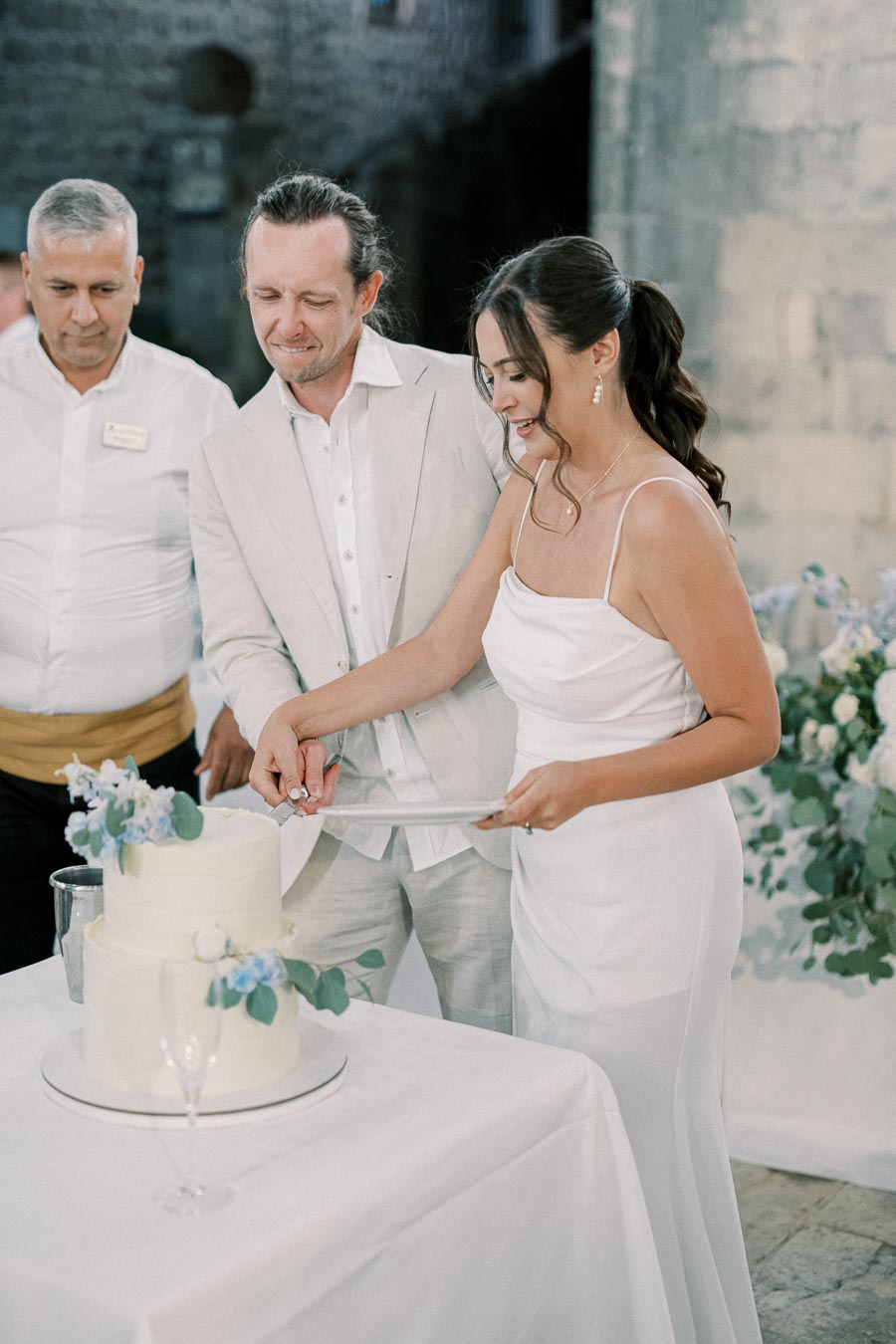 A couple in elegant attire cutting a two-tier wedding cake adorned with blue flowers, surrounded by a romantic outdoor setting.