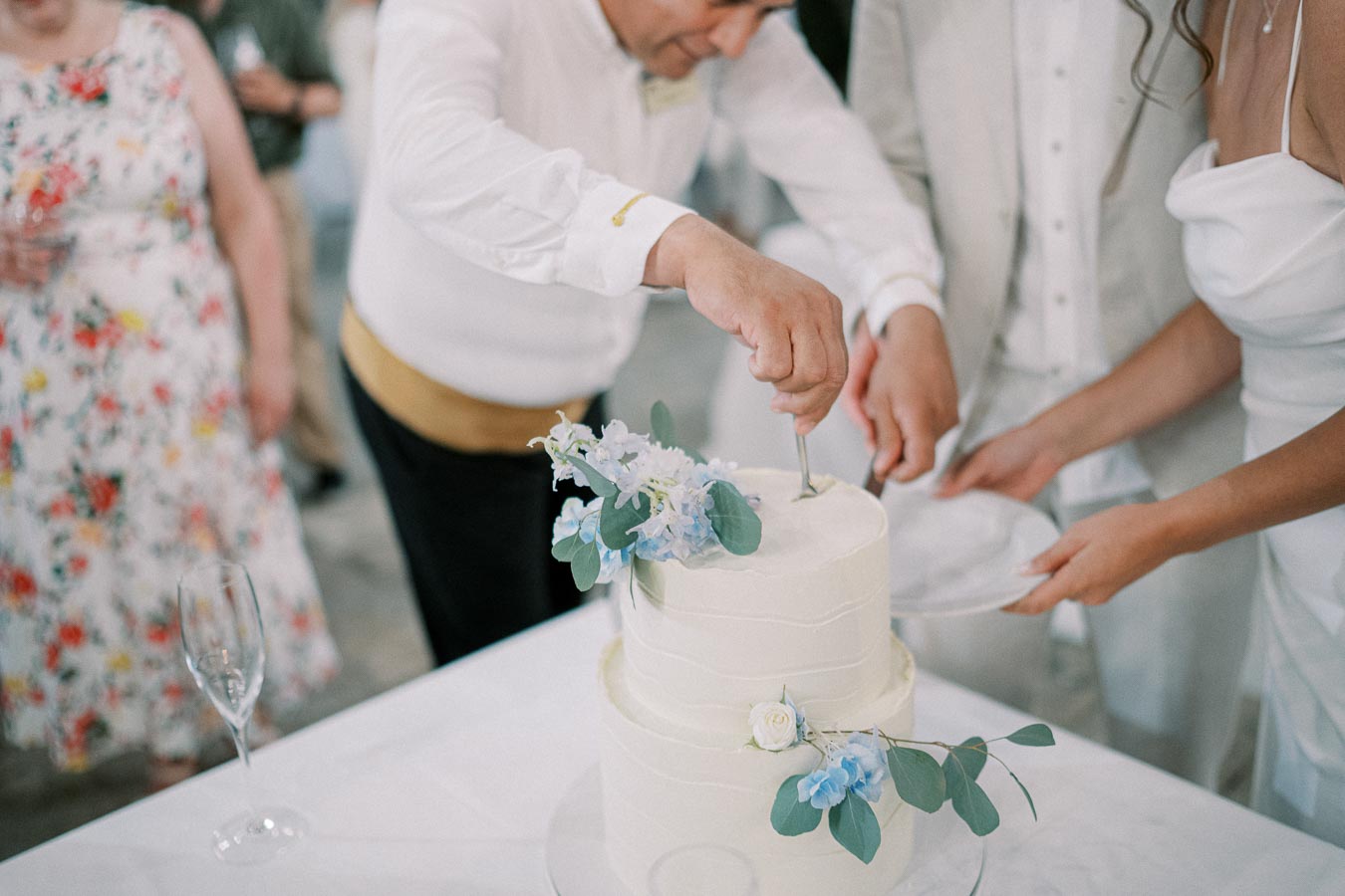 A couple cutting a two-tiered white wedding cake adorned with blue and white flowers, surrounded by guests at a wedding reception.