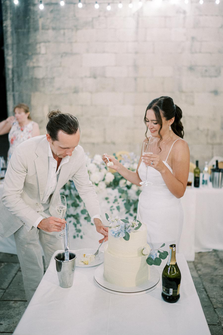 Bride and groom cutting a tiered wedding cake decorated with flowers, celebrating with champagne in an elegant venue.