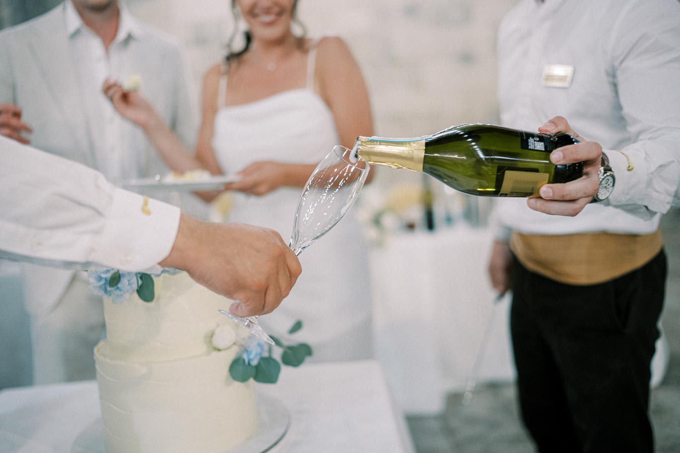 Wedding reception with a close-up of champagne being poured into a glass beside a white tiered wedding cake decorated with blue flowers, with a smiling couple in the background.