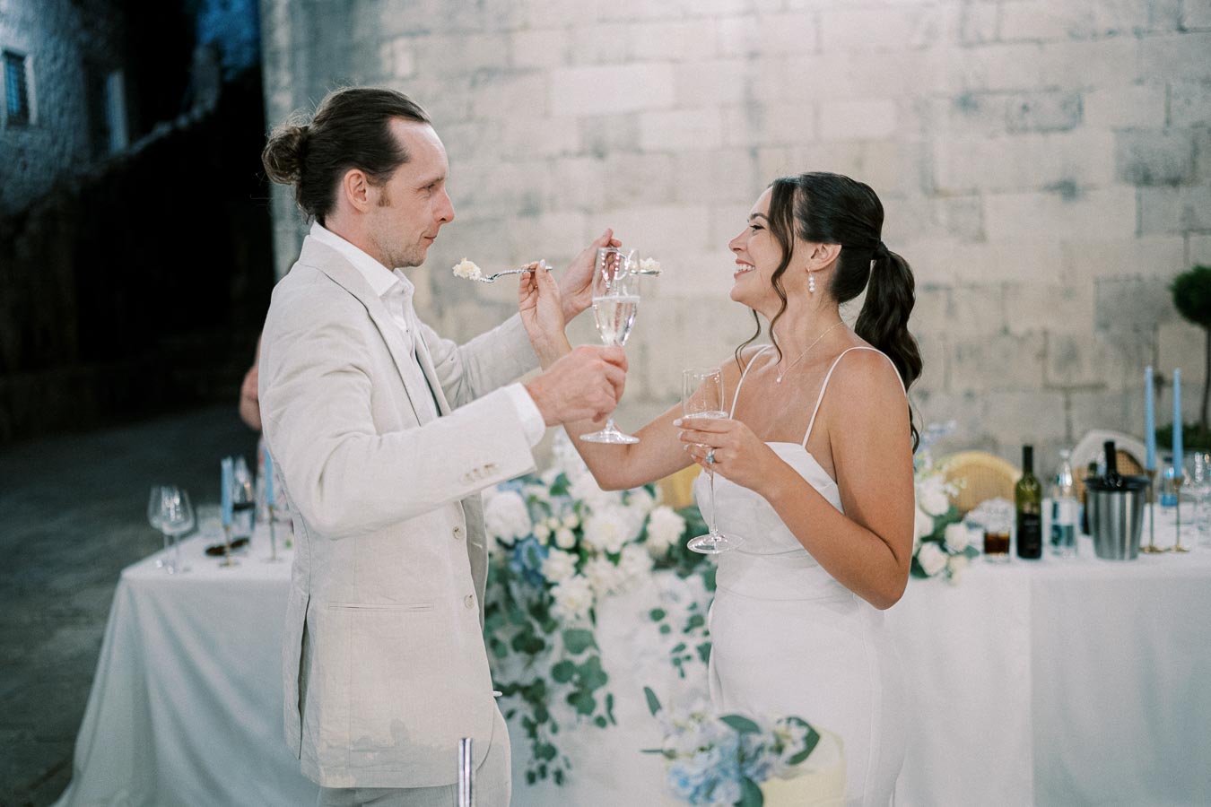 Elegant bride and groom share a joyful champagne toast at their wedding reception, with a beautifully decorated table in the background, creating a romantic atmosphere.
