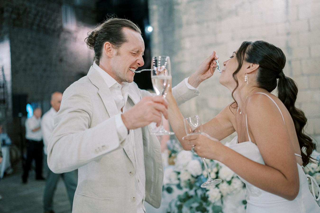 A joyful bride and groom laughing and sharing a treat at their wedding reception, both holding champagne glasses and dressed in elegant attire, symbolizing celebration and happiness.