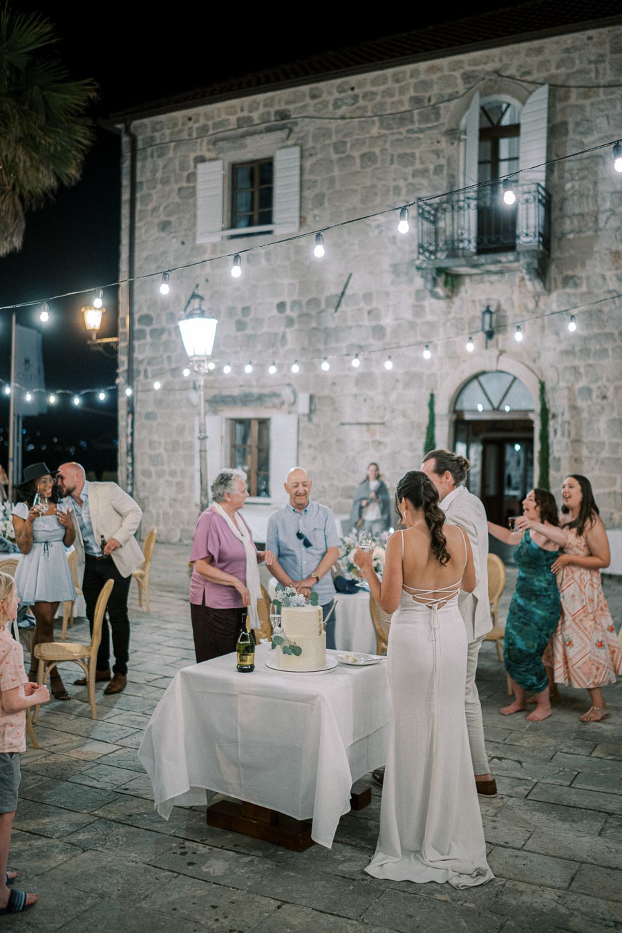 Outdoor evening wedding celebration with guests socializing around a decorated cake table, under string lights near a rustic stone building.