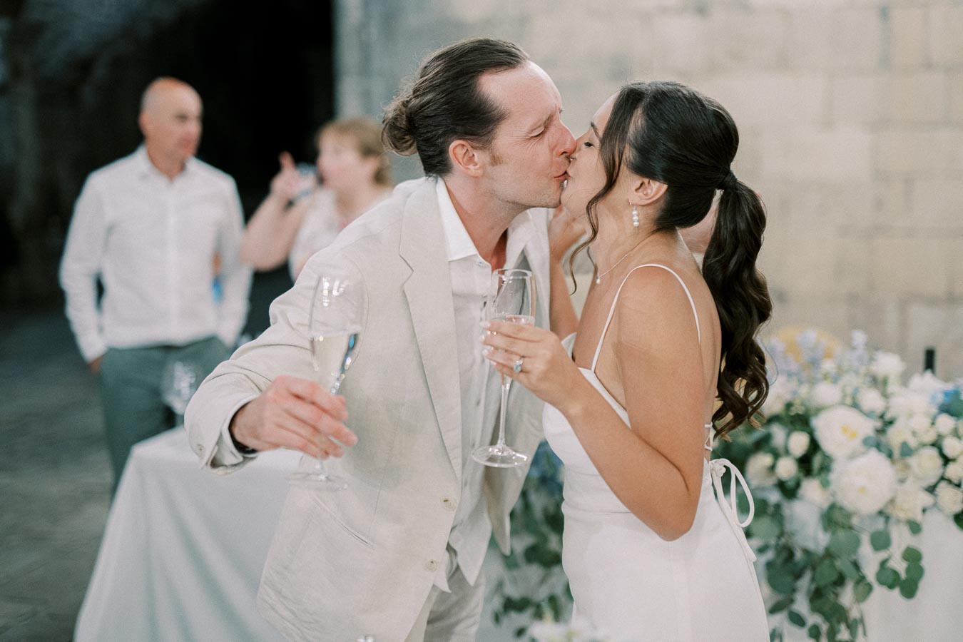 A couple shares a celebratory kiss while holding champagne glasses at a wedding reception, with a backdrop of floral arrangements and guests mingling in the background.