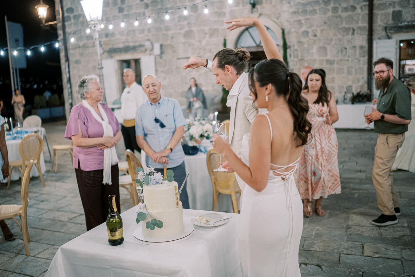 A bride and groom celebrating with guests at an outdoor wedding reception, standing by a decorated cake with champagne, under string lights against a rustic stone building backdrop.