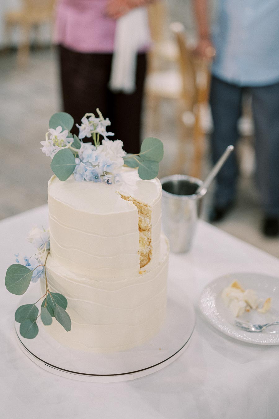 A two-tiered white wedding cake adorned with light blue flowers and eucalyptus leaves, partially sliced, displayed on a white tablecloth with a cake knife and a small plate holding cake pieces nearby.
