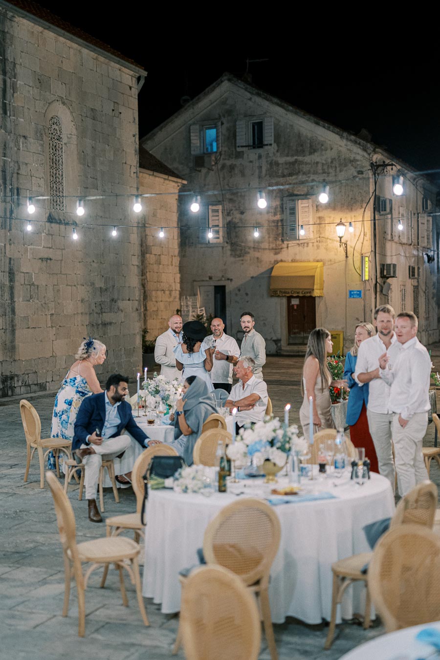 Outdoor evening wedding reception with elegantly dressed guests gathered around tables decorated with white tablecloths and floral centerpieces, set in a historic stone courtyard with string lights illuminating the scene.
