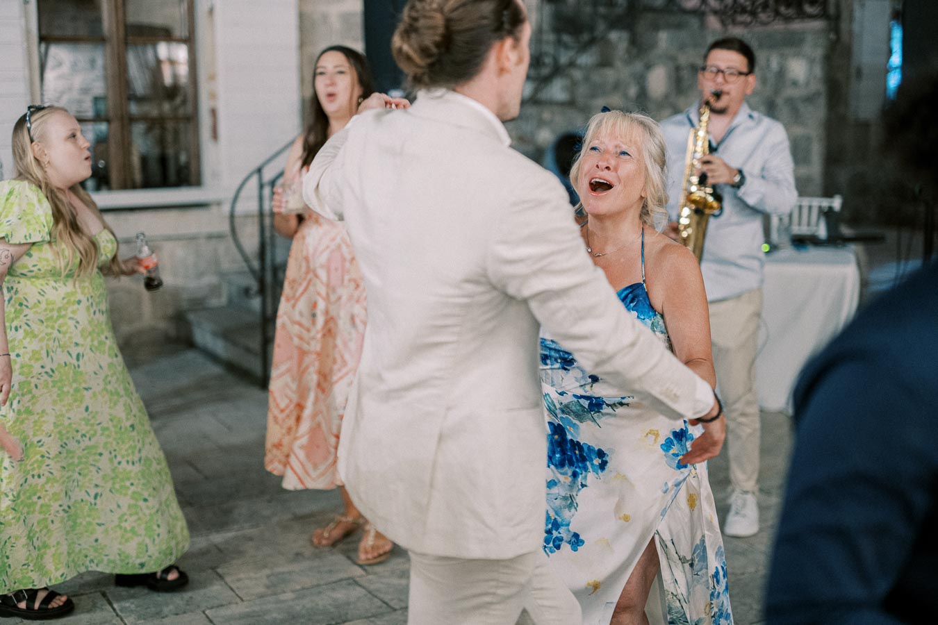 A joyful group of people dancing at an outdoor event, with a live musician playing a saxophone in the background, surrounded by stone architecture.