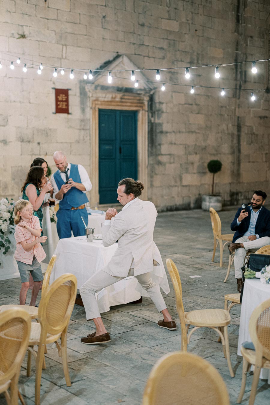 A lively wedding reception scene in an outdoor courtyard, featuring a man in a beige suit dancing energetically with a child. Guests in the background are laughing and capturing the moment on their phones, with festive string lights illuminating the stone walls and blue door.