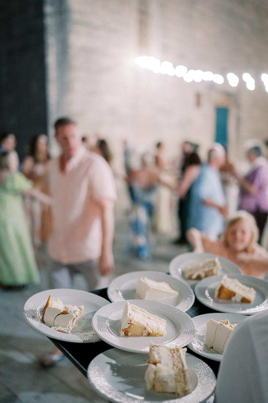 Slices of wedding cake served on plates at an outdoor reception with blurred guests in the background, creating a festive and celebratory atmosphere.