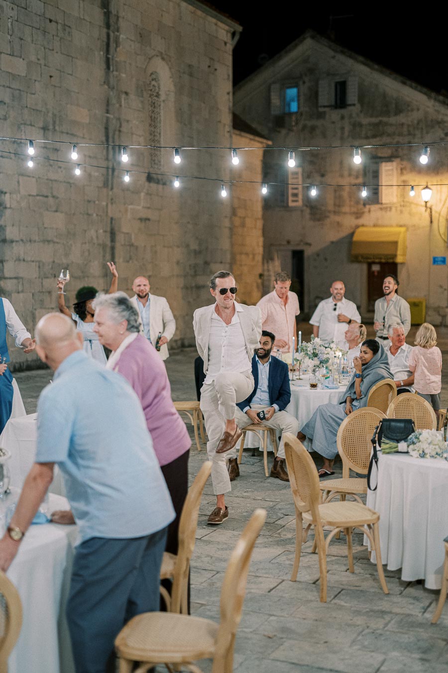 A lively outdoor wedding reception with guests in formal attire enjoying the evening. A man in a light suit and sunglasses dances energetically among tables decorated with flowers, under string lights against a historic stone building.