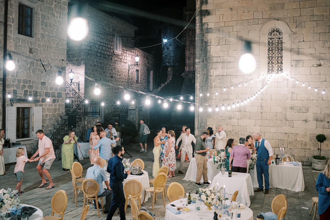Outdoor evening wedding reception in a rustic courtyard, featuring guests dancing and socializing under string lights with stone buildings in the background. Tables are adorned with floral centerpieces, creating a romantic and festive atmosphere.