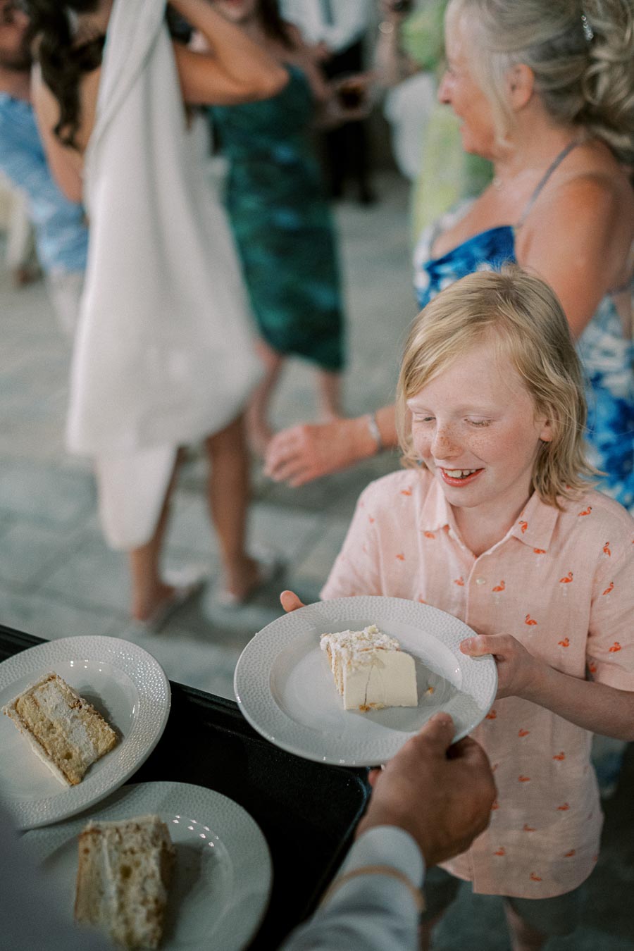 A young boy in a light pink shirt smiling as he receives a slice of cake on a plate during a festive gathering, with blurred figures and additional cake slices in the background.