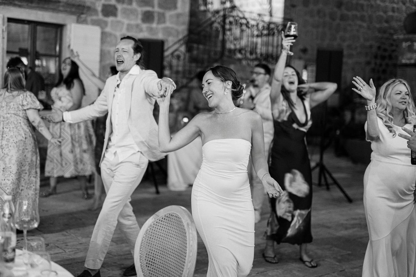 A joyful wedding celebration with guests dancing energetically in a rustic stone venue, featuring a woman in a strapless white dress and a man in a light suit leading the dance.