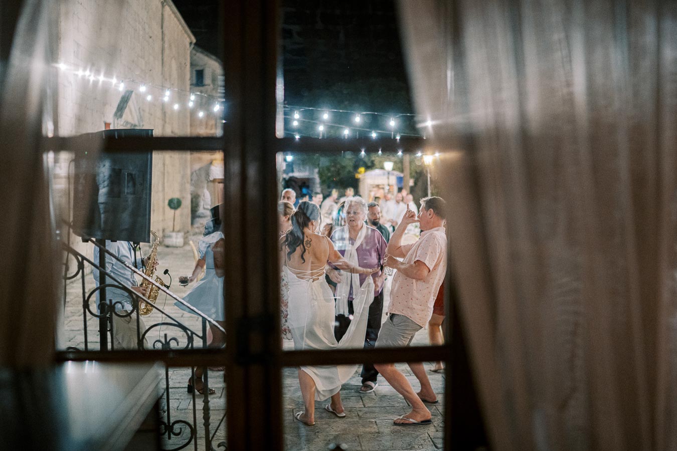 Outdoor wedding reception with guests dancing under string lights, seen through a window frame, creating a festive and joyful atmosphere.