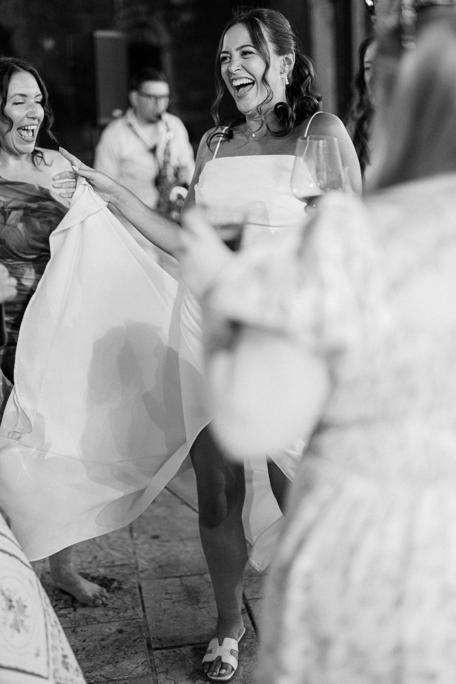 A joyful woman in a white dress laughing and dancing with friends at an indoor celebration, holding a glass of wine. Black and white photography capturing a lively social gathering.