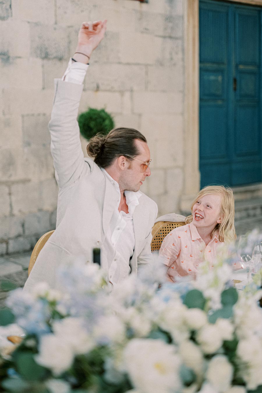 Man in a white suit raising his arm sits at an outdoor table with a smiling child in a pink shirt, a blurred background featuring a stone building and blue door.