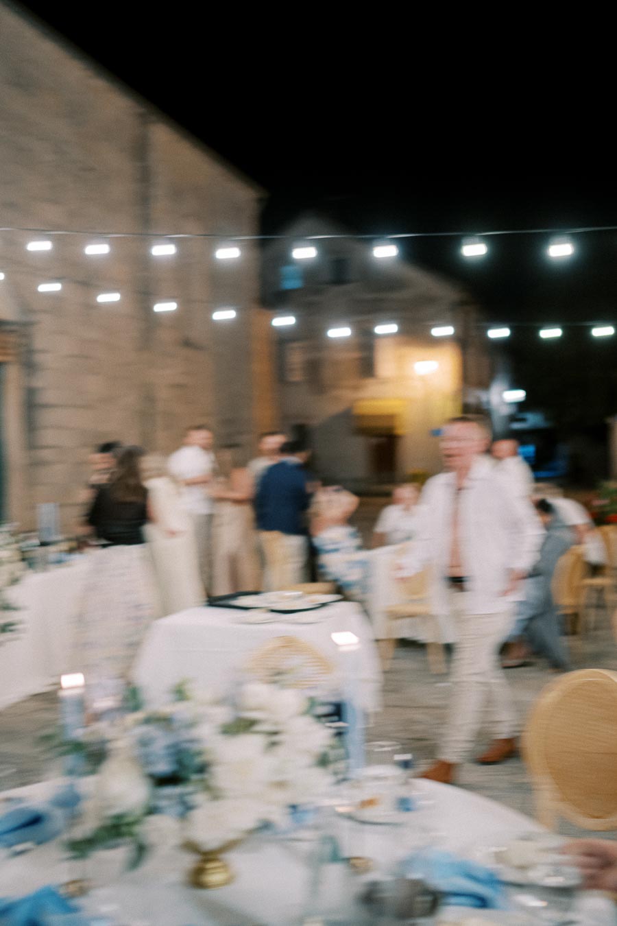Blurred outdoor evening wedding reception scene with string lights, guests mingling, and elegantly decorated tables.