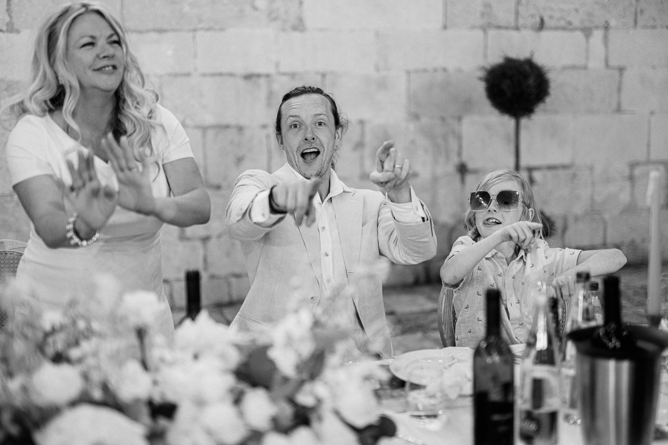 Black and white image of three people joyfully pointing and smiling at a dining table, adorned with flowers and bottles, against a stone wall backdrop.