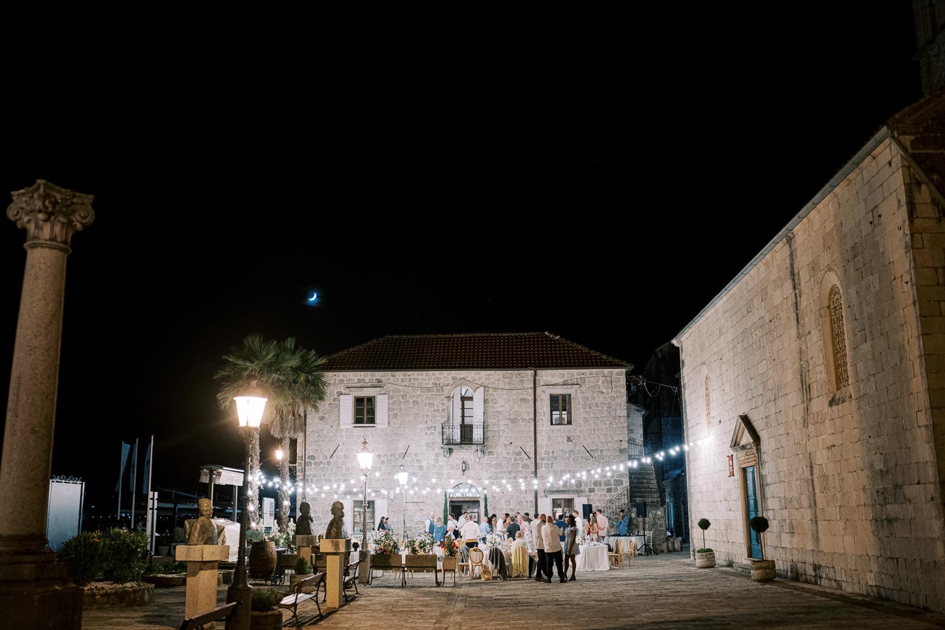 Outdoor evening wedding reception in a historical stone courtyard with string lights, guests mingling under a clear night sky with a crescent moon.