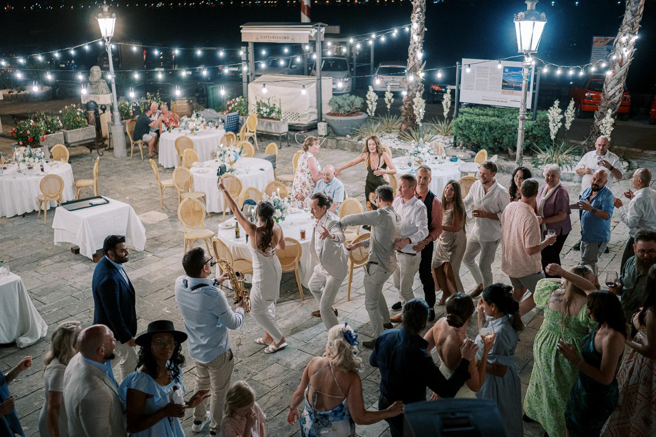 Outdoor evening wedding reception with guests dancing under string lights, surrounded by elegantly decorated tables and vibrant floral arrangements; a saxophonist plays lively music.