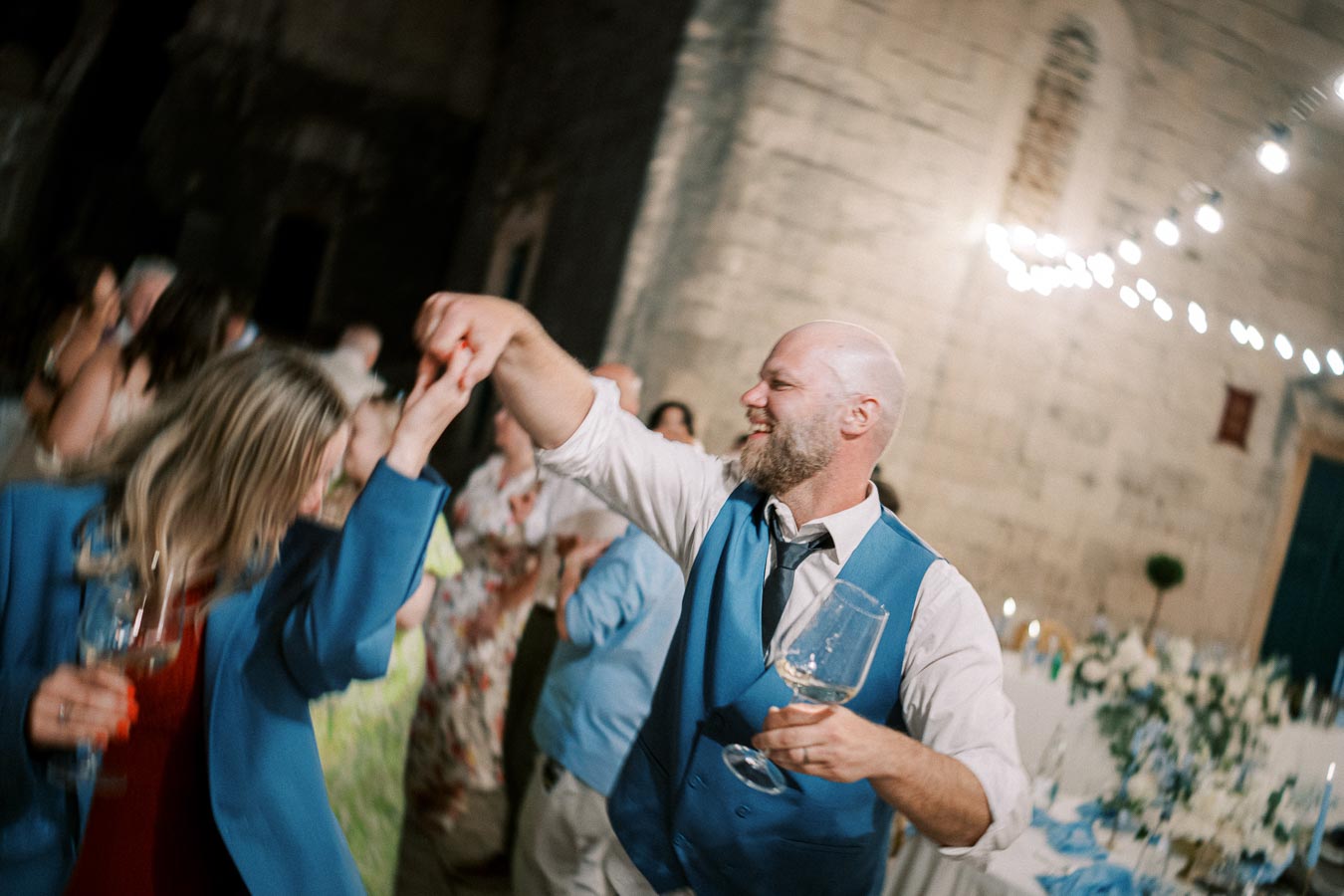 Joyful wedding celebration with a couple dancing under string lights, holding wine glasses, with guests in the background and a historic stone-wall setting.