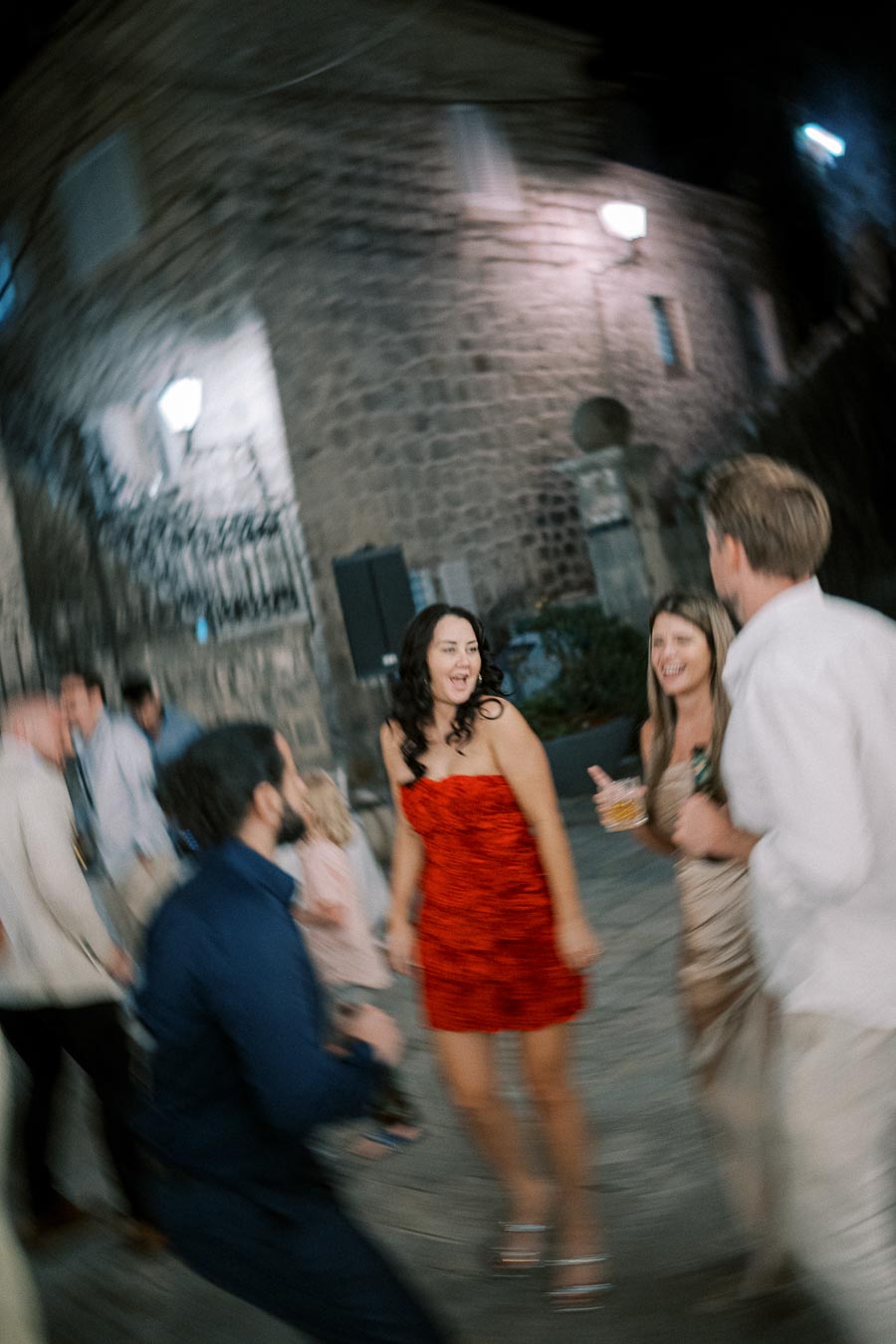 Blurry night scene of a group of people laughing and dancing outdoors, with a stone building illuminated by streetlights in the background.