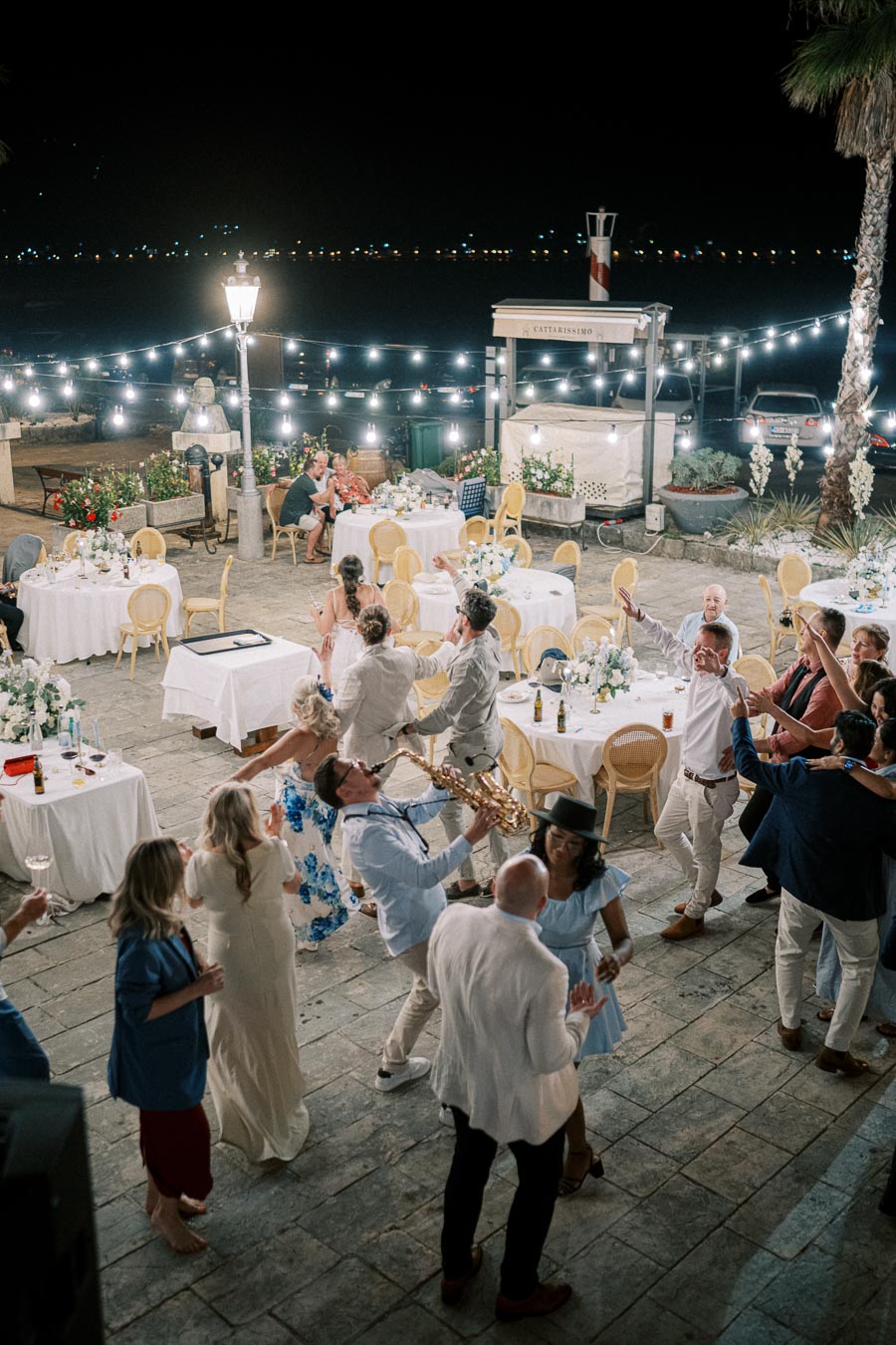 Nighttime outdoor wedding reception with guests dancing and enjoying live saxophone music under string lights. Round tables with floral centerpieces decorate the venue, creating a festive and elegant atmosphere by the water.